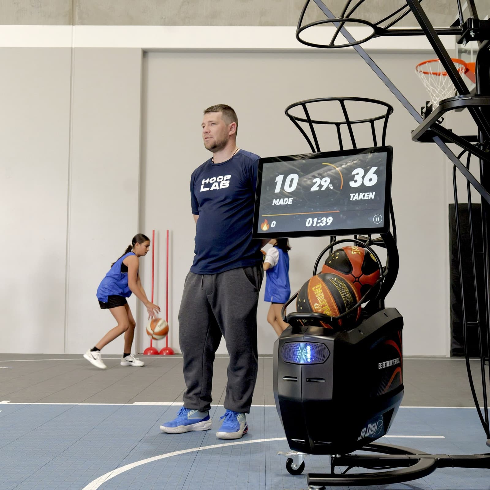 HoopLab coach beside the Dr. Dish CT+ scoreboard while a player shoots in the Shooting Lab, Malaga WA